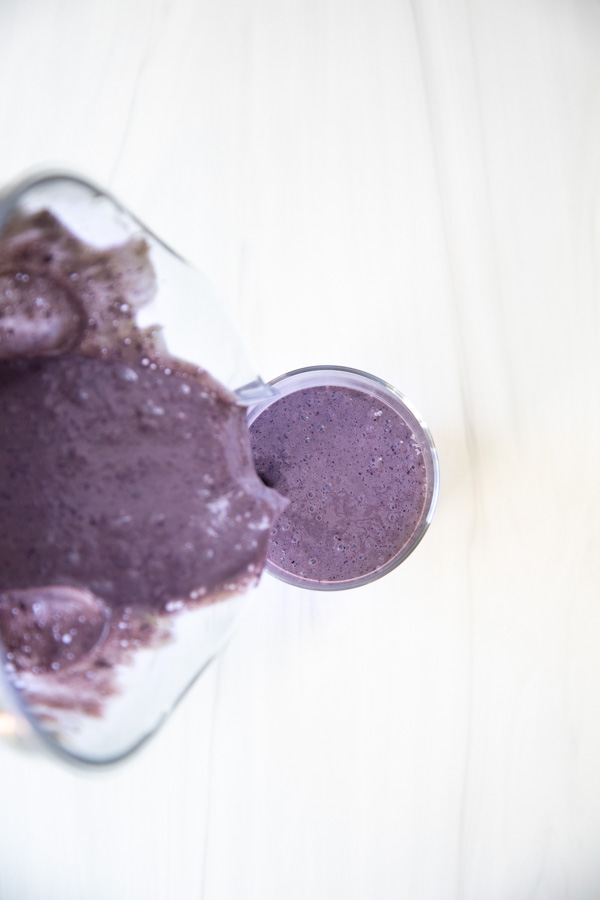 A pitcher of thick purple smoothie is being poured into a glass on a white surface, viewed from above.