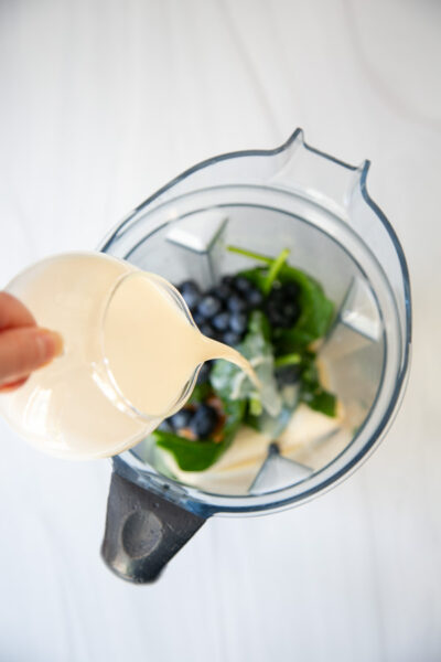 A hand pours creamy liquid from a small pitcher into a blender filled with spinach, blueberries, and other ingredients, preparing to make a smoothie.