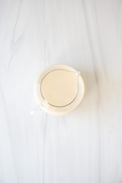 A glass pitcher filled with a light beige liquid sits on a white marble surface, viewed from above.