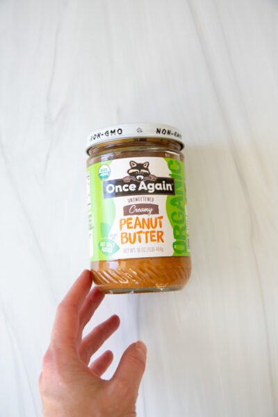 A hand holds a jar of Once Again Organic Creamy Unsweetened Peanut Butter against a white background.