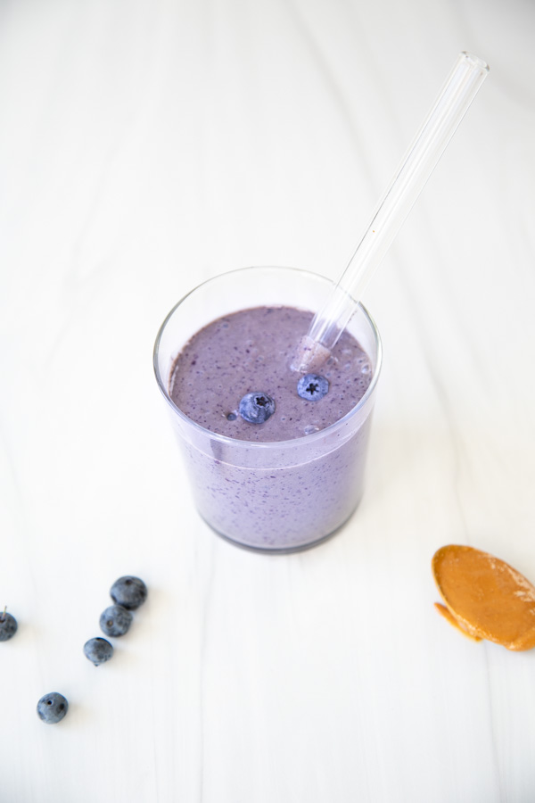 A glass of purple blueberry smoothie with a straw, topped with blueberries. Nearby, there are scattered blueberries and a spoon with peanut butter on a white surface.
