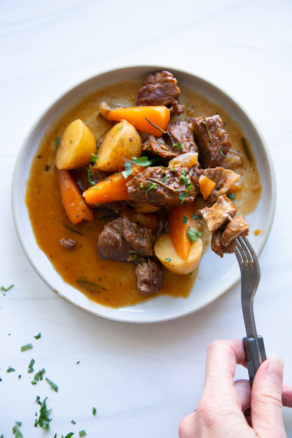 A fork lifts a piece of beef from a plate of beef stew with carrots, potatoes, and herbs.
