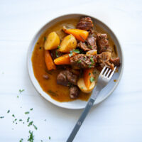 A bowl of beef stew with potatoes and carrots, garnished with herbs and served with a fork.