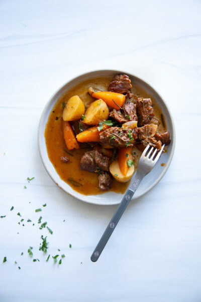 A bowl of beef stew with potatoes and carrots, garnished with herbs and served with a fork.