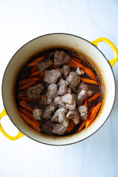 Chunks of browned meat cooking with sliced carrots in a yellow Dutch oven.