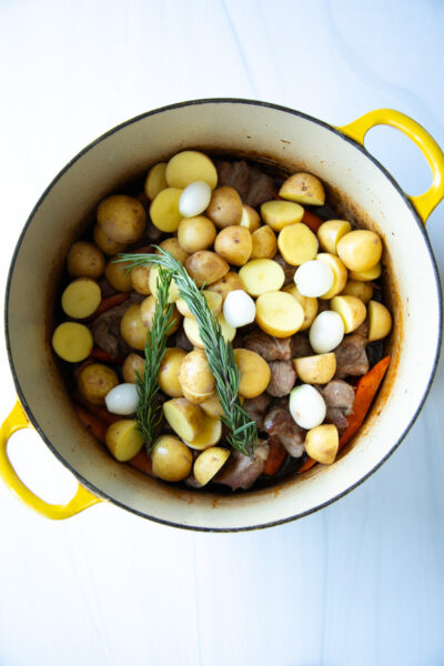 A yellow dutch oven filled with cubed meat, halved potatoes, small onions, carrot pieces, and two sprigs of fresh rosemary.