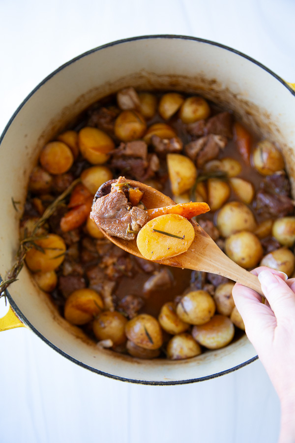 A wooden spoon lifts a piece of meat, a potato slice, and a carrot from a Dutch oven filled with beef stew.