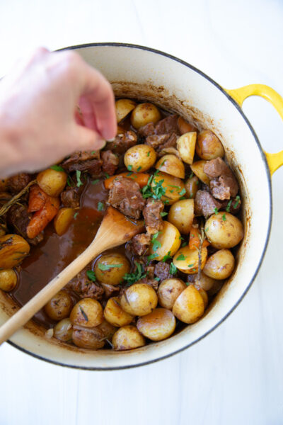 A hand sprinkles fresh herbs over a beef and potato stew in a yellow enameled cast iron pot.