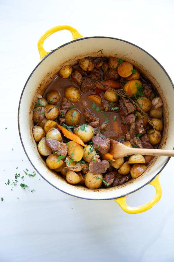 Beef stew with potatoes, carrots, and herbs in a yellow Dutch oven with a wooden spoon.