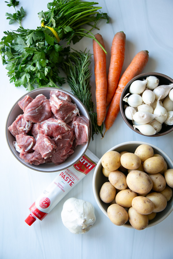 Ingredients for a stew arranged on a white surface, including bowls of raw cubed meat, baby potatoes, and pearl onions, alongside carrots, fresh herbs, a head of garlic, and a tube of tomato paste.